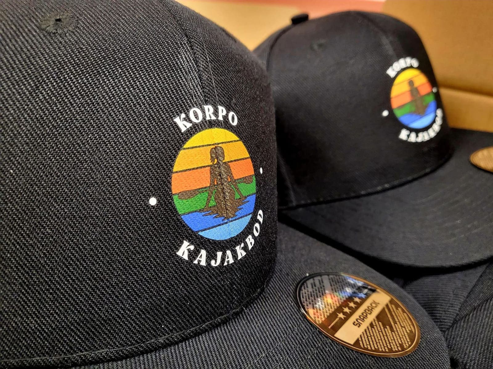 Svarta kepsar med Korpo Kajakbods färgade logotyp