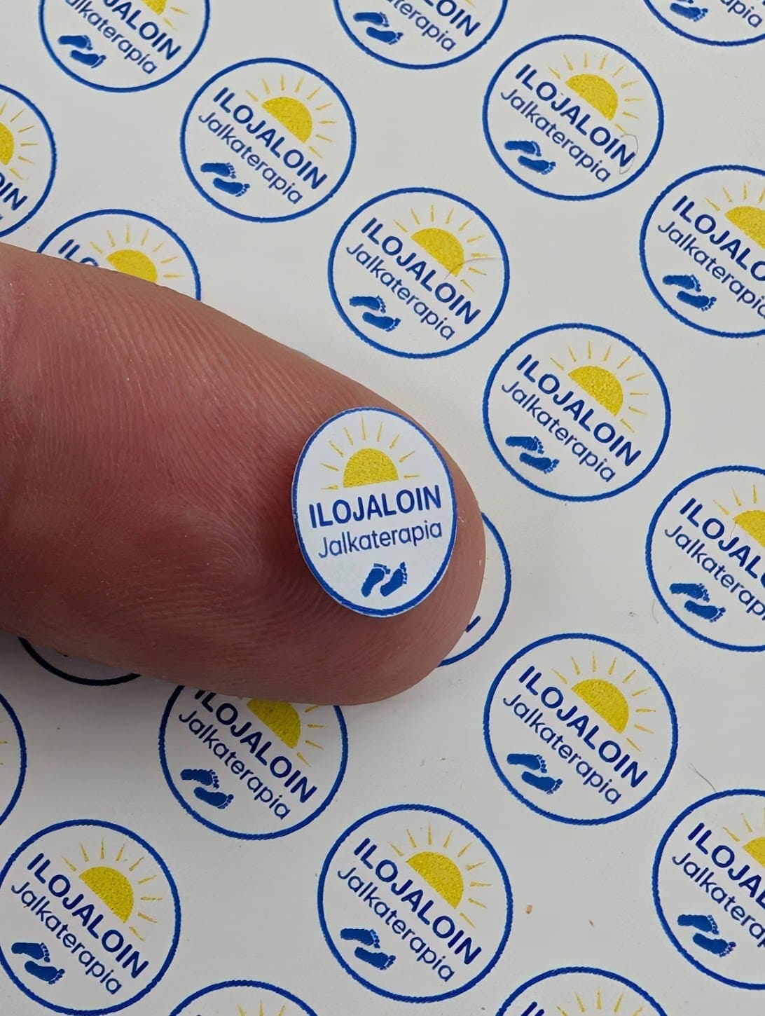 Mycket små runda Ilojaloin-logoetiketter bredvid ett finger